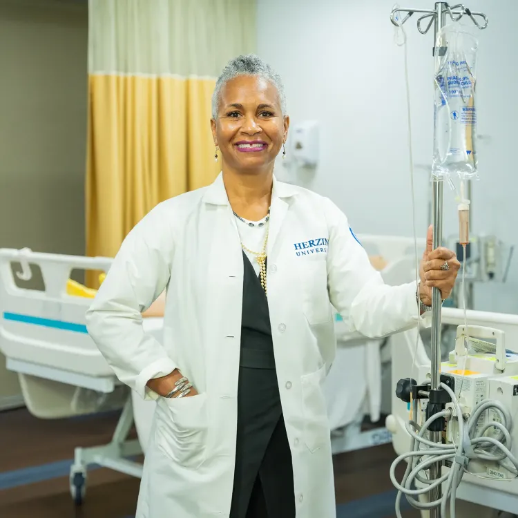 A nursing instructor at Herzing University stands confidently in a medical simulation lab, wearing a white lab coat and smiling while holding an IV pole next to a hospital bed and medical equipment.
